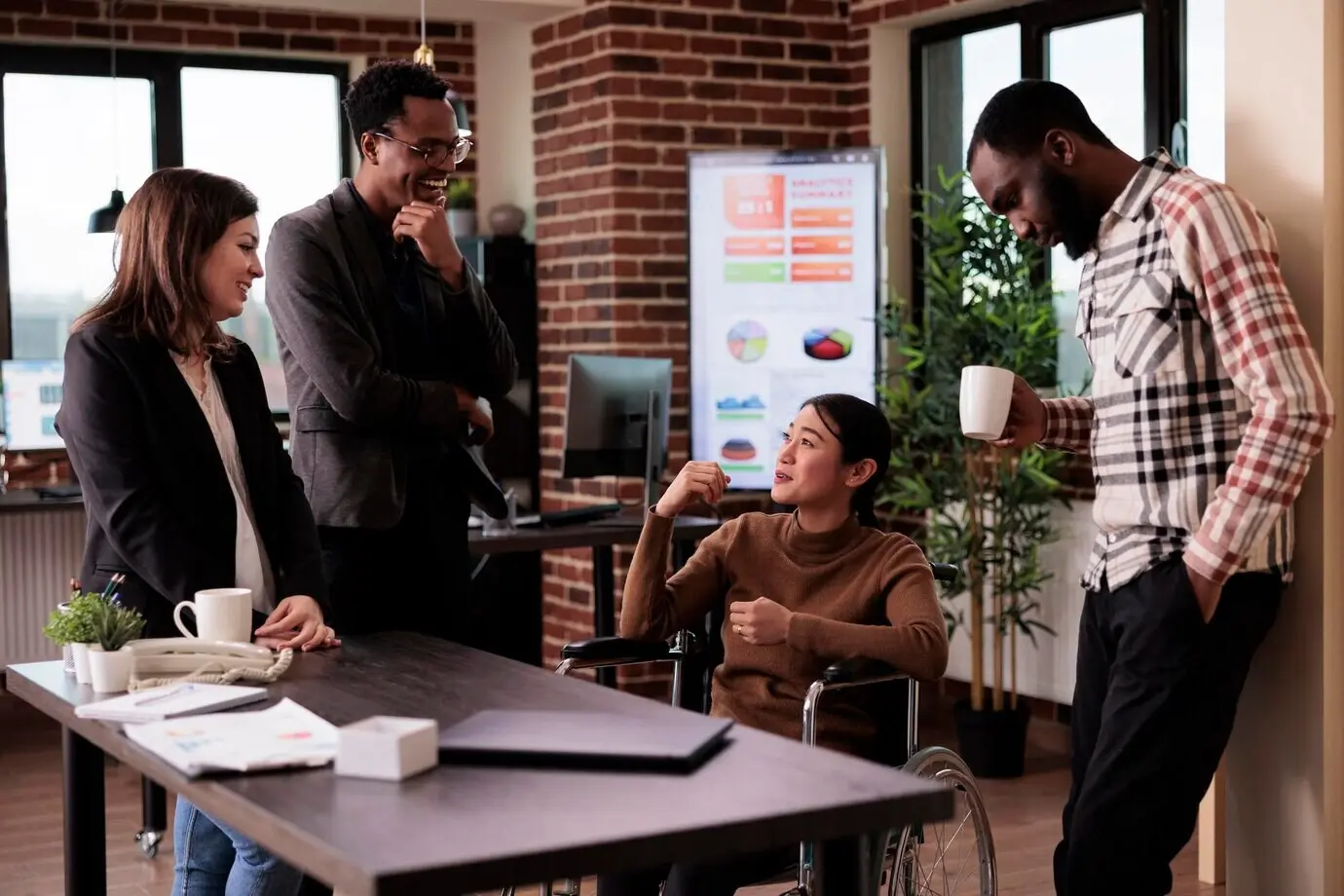 Mujer asiática usuaria de silla de ruedas con discapacidad, riendo con personas en la oficina de una startup durante el descanso de una reunión. Trabajadora que lidia con una condición de salud crónica, hablando con colegas alegres.